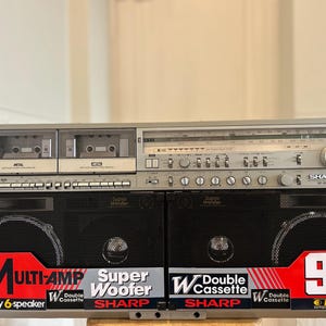 Peut inclure: Un radiocassette Sharp argenté doté d'un double lecteur de cassettes, d'un tuner radio AM/FM et de deux grands haut-parleurs noirs. L'avant présente des étiquettes rouges et blanches indiquant Multi-Amp, Super Woofer, Double Cassette et 90W PMPO.