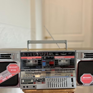 Puede incluir: Un radiocassette Sharp GF-800 plateado con doble pletina de casete, ecualizador gráfico de 10 bandas y asa de transporte superior. La unidad muestra la marca 52W en las rejillas de los altavoces e incluye una antena retráctil y pantalla.