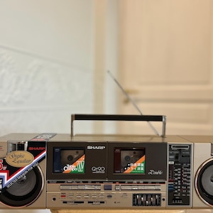 Puede incluir: Un reproductor de casetes doble portátil Sharp QT90 en color plata y negro con asa superior y antena extensible. El frontal cuenta con dos pletinas, sintonizador de radio, controles deslizantes de ecualizador y altavoces con etiquetas 25W y APSS.