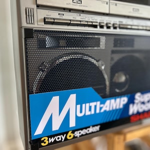 Pu&ograve; includere: Un boombox Sharp argentato con una griglia dell'altoparlante in metallo nero e un'etichetta blu con testo bianco che recita MULTI-AMP Super Woofer e 3way 6-speaker. La sezione superiore include pulsanti di controllo e un lettore cassette.