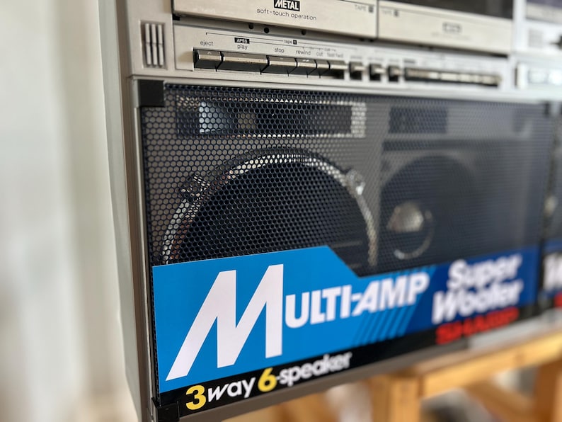 Pu&ograve; includere: Un boombox Sharp argentato con una griglia dell'altoparlante in metallo nero e un'etichetta blu con testo bianco che recita MULTI-AMP Super Woofer e 3way 6-speaker. La sezione superiore include pulsanti di controllo e un lettore cassette.