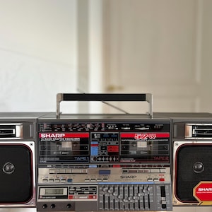 Puede incluir: Un radiocassette Sharp gris con doble pletina, ecualizador gráfico de 10 bandas y dos altavoces grandes. El panel frontal muestra el logotipo de Sharp, la potencia de 52W y varios botones. Un asa de transporte negra está fijada en la parte superior.
