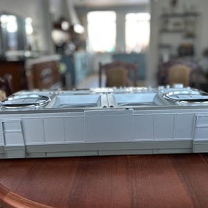 May include: A rectangular silver and grey plastic boombox with two circular speaker grilles on the top surface. The device features a flat, elongated design with vertical panel detailing along the front edge and integrated side latches.