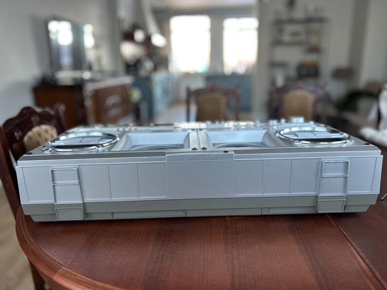 May include: A rectangular silver and grey plastic boombox with two circular speaker grilles on the top surface. The device features a flat, elongated design with vertical panel detailing along the front edge and integrated side latches.