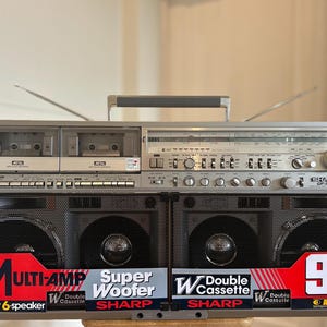 Puede incluir: Un radiocassette Sharp GF-777 plateado con doble pletina, sintonizador de radio y cuatro altavoces frontales. La unidad incluye un asa de transporte, dos antenas extensibles y pegatinas promocionales que dicen Multi-Amp, Super Woofer y 90W.