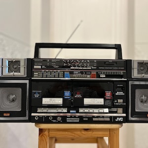 Op de afbeelding: Zwart JVC draagbaar componentensysteem met zilveren accenten, met radio, cassettespelers en luidsprekers. De boombox heeft een handvat en staat op een houten kruk.