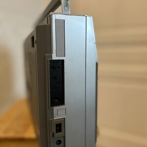 May include: Silver vintage boombox with a retractable handle. The side panel features a speaker grill, input jacks, and a power port. The boombox is set on a wooden stool.