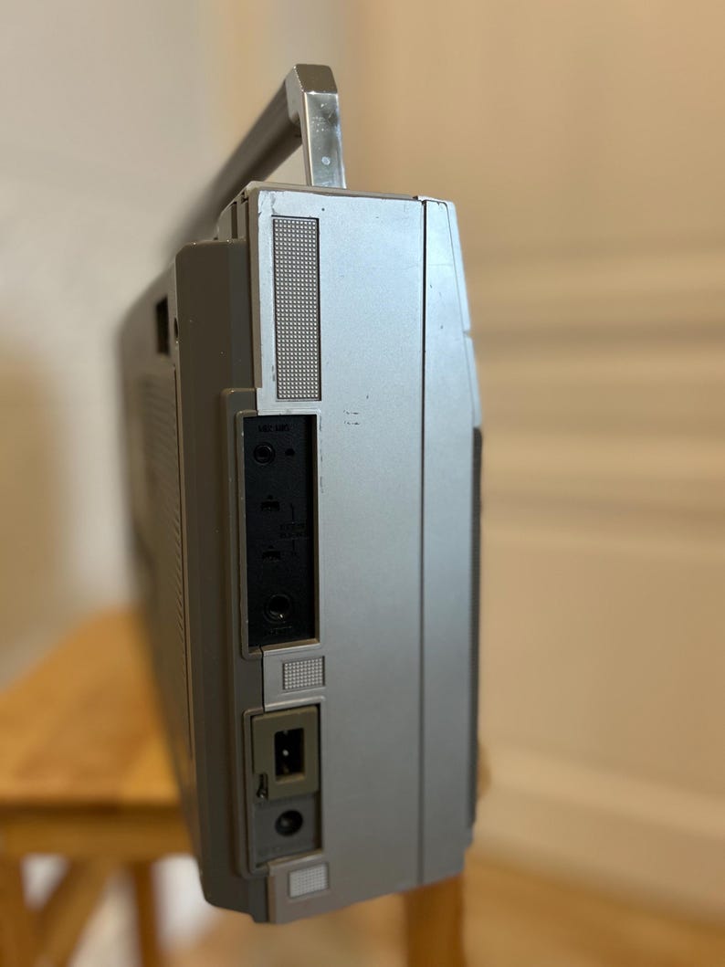 May include: Silver vintage boombox with a retractable handle. The side panel features a speaker grill, input jacks, and a power port. The boombox is set on a wooden stool.