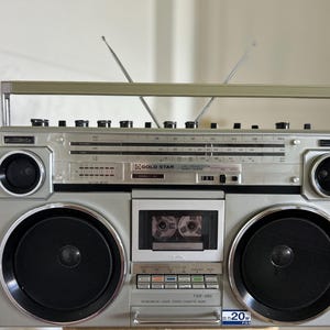 Może przedstawiać: Srebrny, przenośny radiomagnetofon stereo Gold Star TSR-580 wyposażony w odtwarzacz kaset, dwa duże głośniki, dwa małe głośniki wysokotonowe, tuner radiowy ze skalą częstotliwości, uchwyt do przenoszenia oraz dwie wysuwane metalowe anteny.