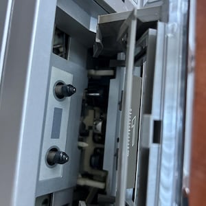Pu&ograve; includere: Un lettore di cassette argentato con lo scomparto per la cassetta aperto. L'interno presenta due perni neri, una testina magnetica e uno sportello in plastica trasparente con una freccia bianca e la scritta APSS sul telaio superiore.