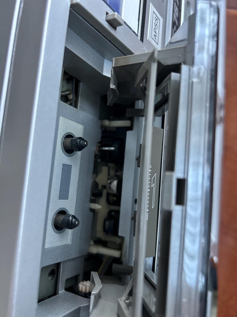 Pu&ograve; includere: Un lettore di cassette argentato con lo scomparto per la cassetta aperto. L'interno presenta due perni neri, una testina magnetica e uno sportello in plastica trasparente con una freccia bianca e la scritta APSS sul telaio superiore.
