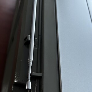 May include: A black electronic device featuring a long, thin, silver-coloured telescopic antenna that is retracted and resting in a moulded groove along the side of the unit. The device has a smooth, matte black finish and a light grey panel section.