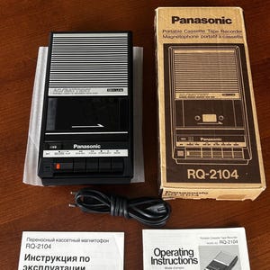 May include: A black and silver Panasonic RQ-2104 portable cassette tape recorder with a matching cardboard box, a black power cord, and two instruction manuals. The device features a speaker grille, control buttons, and the Panasonic logo.