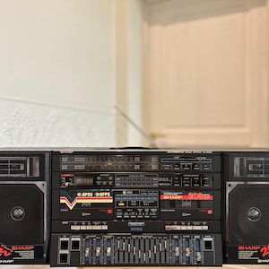 Könnte beinhalten: Ein schwarzer Sharp HK-9000 Ghettoblaster mit zwei Kassettendecks, einem Radiotuner und einem grafischen Equalizer. Die Frontplatte zeigt den Text Sharp 208W PMPO HK-9000 in roten und gelben Akzenten, mit zwei großen Lautsprechern an den Seiten.