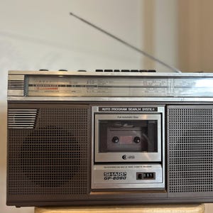 May include: A brown and silver Sharp GF-8060 radio cassette recorder featuring a top-mounted extendable antenna, a horizontal radio frequency tuning display, dual speakers, and a central cassette deck labeled Auto Program Search System.