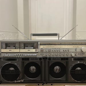 May include: Silver vintage boombox with dual cassette decks, radio dial, and large speakers. The boombox has two antennas and a handle for portability. The speakers have a mesh grill design. The brand name "SHARP" is visible.