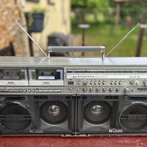 May include: A silver Sharp GF-777 boombox featuring a double cassette deck, a radio tuner with a frequency display, and four front-facing speakers protected by black metal grilles. The portable audio system includes a top handle and two antennas.