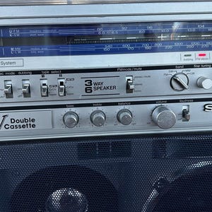 Pu&ograve; includere: Un registratore radio a doppia cassetta stereo Sharp GF-767 color argento con un display blu per la frequenza radio, diverse manopole di controllo, interruttori per funzione, livello di registrazione e selezione nastro, e una griglia nera.