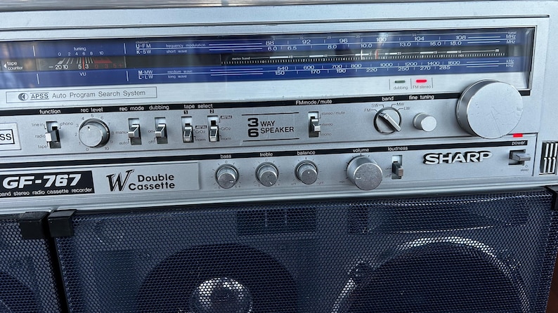 Pu&ograve; includere: Un registratore radio a doppia cassetta stereo Sharp GF-767 color argento con un display blu per la frequenza radio, diverse manopole di controllo, interruttori per funzione, livello di registrazione e selezione nastro, e una griglia nera.