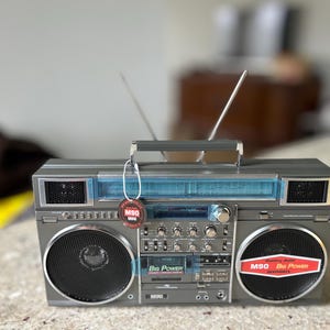 May include: A grey retro boombox with a handle, aerials, and speakers. The boombox has a blue tinted plastic panel and a tag that says "M90 Mini". The speakers have red and white labels that say "M90 Big Power".