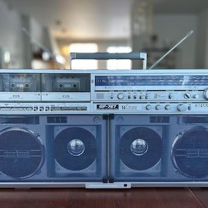 May include: A silver and black Sharp GF-787 boombox with a handle, antenna, and cassette decks. The speakers are covered with a blue mesh. The boombox has a radio tuner and various control knobs and buttons. The text "GF-787 W Double Cassette" is visible.