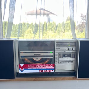 Könnte beinhalten: Eine silberne Sharp Stereoanlage mit vertikalem Plattenspieler mit Linear-Tracking-Tonarm, Kassettendeck und Radiotuner. Das Gerät umfasst zwei abnehmbare schwarze Lautsprecher und ein Frontetikett mit der Aufschrift BSLT BOTH SIDES PLAY.