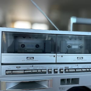Pu&ograve; includere: Un lettore di cassette portatile color argento con doppio vano per cassette etichettato Tape 1 e Tape 2. Ogni vano include uno sportello in plastica trasparente e la scritta Metal soft-touch operation. Sotto si trova una fila di tasti.