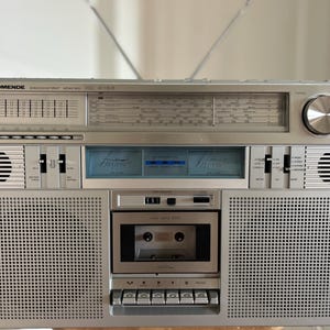 Könnte beinhalten: Ein silberner Nordmende Discocorder Stereo 4155 Ghettoblaster mit Radiotuner und Frequenzskala, Kassettendeck mit mechanischen Tasten, zwei integrierten Lautsprechern, einer zentralen blau beleuchteten Pegelanzeige und einer Teleskopantenne.