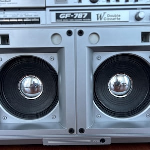 Pu&ograve; includere: Un registratore a doppia cassetta Sharp GF-767 color argento con due grandi altoparlanti neri dotati di parapolvere cromati. Il pannello frontale mostra il nome del modello GF-767 e la scritta W Double Cassette, oltre a varie manopole.