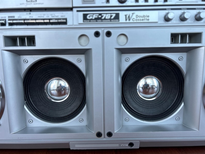 Pu&ograve; includere: Un registratore a doppia cassetta Sharp GF-767 color argento con due grandi altoparlanti neri dotati di parapolvere cromati. Il pannello frontale mostra il nome del modello GF-767 e la scritta W Double Cassette, oltre a varie manopole.