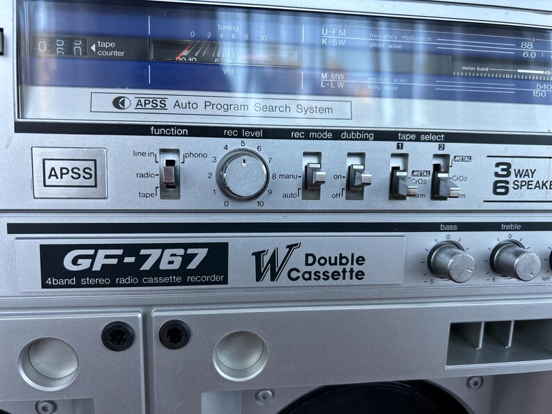 May include: A silver GF-767 4-band stereo radio cassette recorder with a double cassette deck. The front panel features an APSS Auto Program Search System, a tuning display, a recording level knob, and various switches for function, mode, and tape selection.