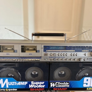 Może przedstawiać: Srebrny przenośny system stereo Sharp z podwójnym magnetofonem kasetowym, tunerem radiowym AM/FM oraz dwoma odłączanymi głośnikami z czarnymi metalowymi maskownicami. Z przodu widnieje napis Multi-Amp, Super Woofer, Double Cassette oraz 90W PMPO.