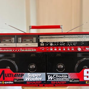 May include: A red Sharp GF-777 boombox featuring a double cassette deck, a radio tuner with a silver dial, and two extendable antennas. The front displays black speaker grilles with labels including Multi-Amp, Super Woofer, and 90W PMPO.