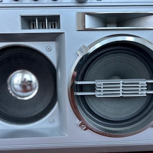 May include: A silver boombox featuring two prominent circular speakers. The left speaker has a smooth metallic centre dome, while the right speaker is framed by a chrome ring and includes a horizontal silver grille across the centre cone.