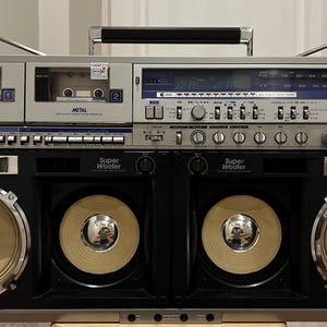 Könnte beinhalten: Ein silber-schwarzer Sharp GF-1000 Ghettoblaster mit zwei Kassettendecks, einem Radiotuner mit blauem Display und vier großen Lautsprechern mit der Aufschrift Super Woofer. Das Gerät verfügt über einen Tragegriff, zwei Antennen und Regler.