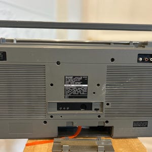 May include: A grey vintage boombox with a silver handle and antenna. The back of the radio has various input and output ports, along with a label that includes the model number and other information. The radio is sitting on a wooden surface.