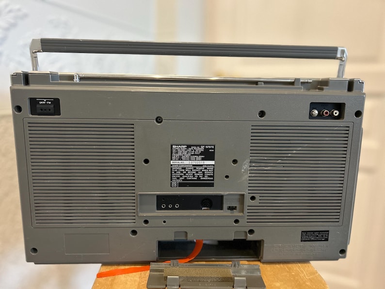 May include: A grey vintage boombox with a silver handle and antenna. The back of the radio has various input and output ports, along with a label that includes the model number and other information. The radio is sitting on a wooden surface.