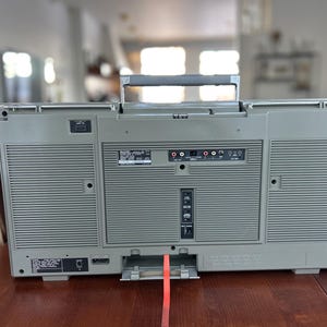 May include: The back view of a grey portable boombox featuring a top carrying handle, two large speaker grilles, and a central panel with various input and output ports, including RCA jacks, a power cord socket, and a vertical switch control panel.