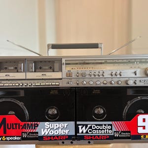 Puede incluir: Un radiocasete estéreo portátil Sharp GF-800 plateado con doble pletina, sintonizador de radio y dos altavoces negros desmontables. El frontal muestra texto que incluye Multi-Amp, Super Woofer, Double Cassette y 90W PMPO.