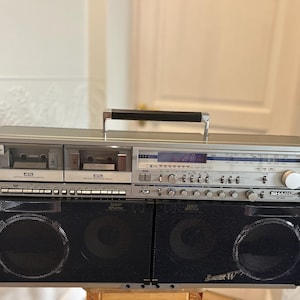 May include: A silver Sharp GF-9696 boombox featuring dual cassette decks, a radio tuner with a digital display, and two large black mesh speaker grilles. The portable audio system includes a black carrying handle and various control knobs.