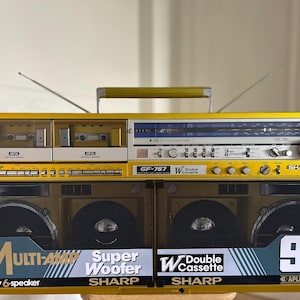 Może przedstawiać: Jaskrawożółty boombox Sharp GF-767 z podwójnym magnetofonem kasetowym, tunerem radiowym z niebieskim wyświetlaczem oraz czterema głośnikami przednimi za czarnymi metalowymi maskownicami. Urządzenie posiada górny uchwyt, dwie anteny i napisy.