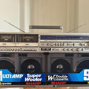May include: A silver Sharp GF-767 boombox featuring a double cassette deck, radio tuner, and two large speaker grilles. The front displays blue labels with text including Multi-Amp, Super Woofer, Double Cassette, and 90W PMPO. It has a top handle.