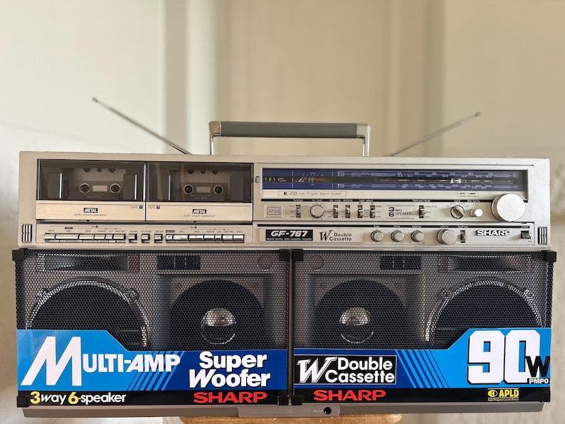 May include: A silver Sharp GF-767 boombox featuring a double cassette deck, radio tuner, and two large speaker grilles. The front displays blue labels with text including Multi-Amp, Super Woofer, Double Cassette, and 90W PMPO. It has a top handle.