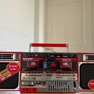 Könnte beinhalten: Ein rot-silberner Sharp GF-800 Ghettoblaster mit zwei Kassettendecks, einem 10-Band-Grafik-Equalizer und zwei abnehmbaren Lautsprechern. Die Frontplatte zeigt das Sharp-Logo, die 52W-Leistungsangabe sowie diverse Tasten und Schieberegler.