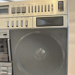 May include: Silver vintage boombox with a mesh speaker cover and various buttons and dials. Features include FM mode, radio memory, and a digital clock display. The boombox has a cassette player and a handle for portability.