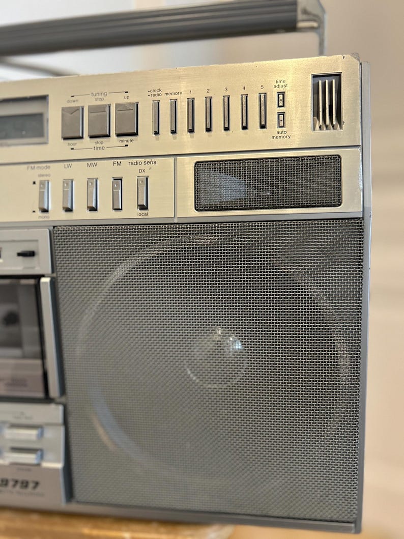 May include: Silver vintage boombox with a mesh speaker cover and various buttons and dials. Features include FM mode, radio memory, and a digital clock display. The boombox has a cassette player and a handle for portability.