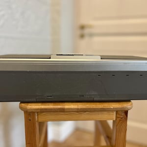 May include: A vintage grey and silver electronic device with a rectangular shape, resting on a small wooden stool. The device has a series of small rectangular openings on the front and a silver top. The stool is made of light-coloured wood.