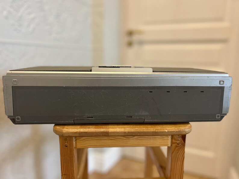 May include: A vintage grey and silver electronic device with a rectangular shape, resting on a small wooden stool. The device has a series of small rectangular openings on the front and a silver top. The stool is made of light-coloured wood.