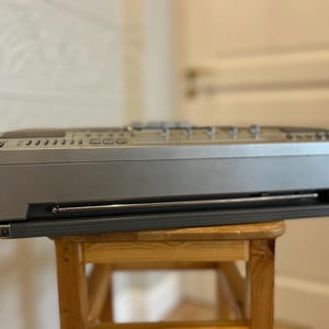 May include: A silver and black vintage boombox with a retractable aerial and control buttons. The boombox is resting on a small wooden stool. The background is a blurred interior.