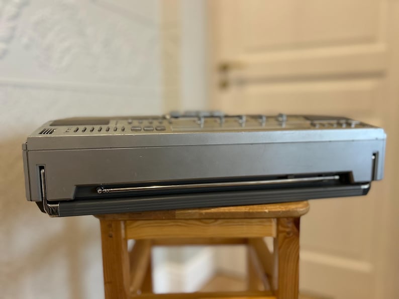 May include: A silver and black vintage boombox with a retractable aerial and control buttons. The boombox is resting on a small wooden stool. The background is a blurred interior.